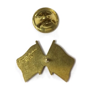 Puede incluir: Vista posterior de un pin de metal dorado con dos banderas cruzadas. El pin tiene un cierre de seguridad en la parte posterior y el texto "Prentice Medals USA" est&aacute; grabado en la parte posterior de una de las banderas.