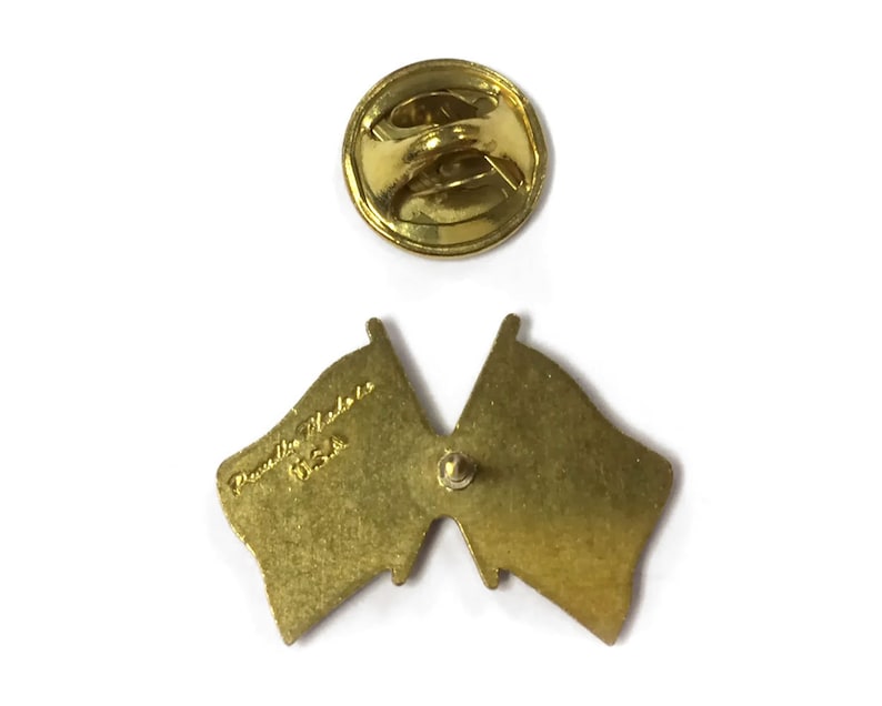 Puede incluir: Vista posterior de un pin de metal dorado con dos banderas cruzadas. El pin tiene un cierre de seguridad en la parte posterior y el texto "Prentice Medals USA" est&aacute; grabado en la parte posterior de una de las banderas.