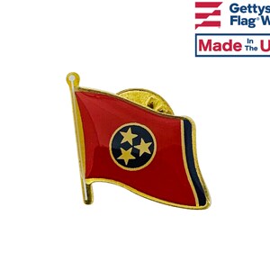 May include: Gold-colored pin with a red, white, and blue Tennessee state flag design. The flag features three white stars on a blue circle in the center. The pin is made in the USA.