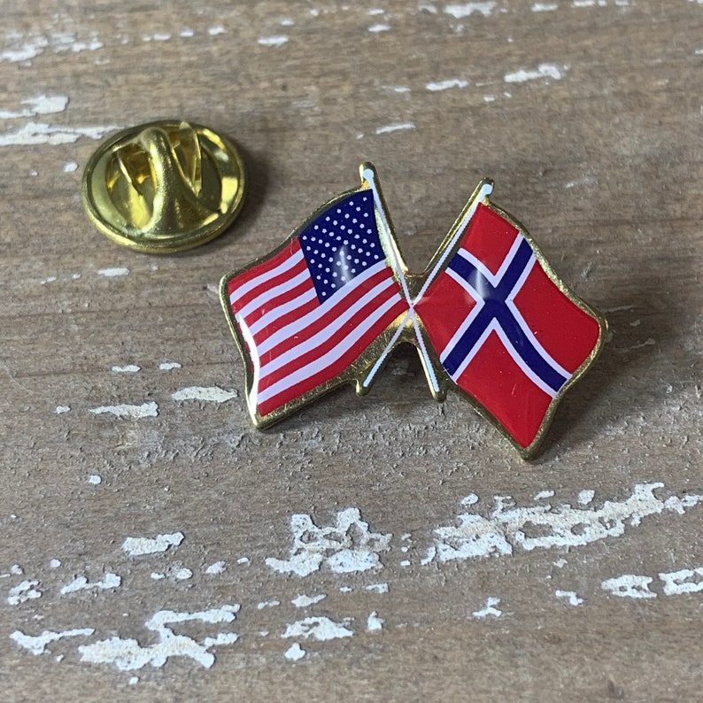 May include: A gold pin with an American flag and a Norwegian flag crossed over each other. The flags are red, white, and blue.