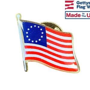 May include: A gold-colored pin featuring a replica of the Betsy Ross flag, the first American flag with 13 stars in a circle on a blue field, and 13 alternating red and white stripes. The pin is made in the USA.
