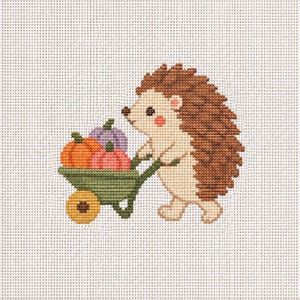 Peut inclure: Un motif de point de croix sur tissu à grille blanc représentant un hérisson brun clair avec des piquants brun foncé poussant une brouette verte. La brouette contient trois petites citrouilles orange, rose et violet clair.