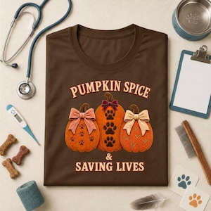 Puede incluir: Una camiseta marrón con un gráfico de tres calabazas naranjas con lazos y huellas, con el texto PUMPKIN SPICE & SAVING LIVES. La camiseta está rodeada por un estetoscopio, un termómetro, un portapapeles, un cuenco y premios para mascotas.