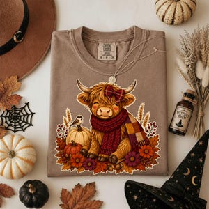 Peut inclure: Un t-shirt marron avec une illustration de vache Highland portant un n&oelig;ud &eacute;cossais rouge et une &eacute;charpe en tricot &eacute;pais. La vache est entour&eacute;e de citrouilles, de feuilles d'automne, d'&eacute;pis de bl&eacute; et d'un petit oiseau. Col rond et &eacute;tiquette Comfort Colors.