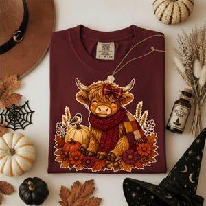 May include: A maroon t-shirt featuring a graphic of a fluffy highland cow wearing a red knit scarf and a plaid bow. The cow is surrounded by autumn leaves, pumpkins, wheat stalks, and flowers. The shirt has a Comfort Colors brand label inside.