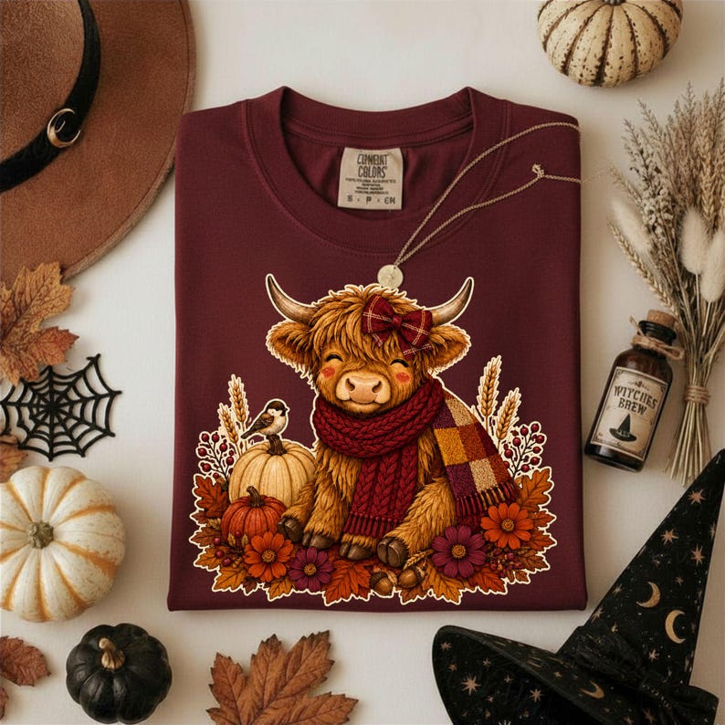 Peut inclure: Un t-shirt marron avec une illustration d'une vache highland duveteuse portant une &eacute;charpe en tricot rouge et un n&oelig;ud &agrave; carreaux. La vache est entour&eacute;e de feuilles d'automne, de citrouilles, d'&eacute;pis de bl&eacute; et de fleurs. &Eacute;tiquette Comfort Colors.