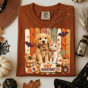 Puede incluir: Una camiseta color óxido con un gráfico de un cachorro golden retriever con pajarita de cuadros y un gatito atigrado con una etiqueta de huella. El diseño incluye calabazas, murciélagos, estrellas y un letrero que dice VET ASSISTANT.