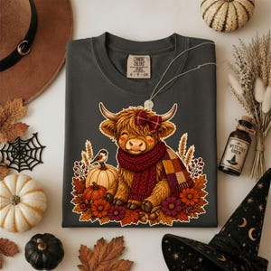 Peut inclure: Un t-shirt gris fonc&eacute; avec une illustration d'une vache Highland poilue portant un n&oelig;ud &agrave; carreaux rouge et une &eacute;charpe en tricot. La vache est entour&eacute;e de citrouilles, de feuilles d'automne, d'un petit oiseau et de fleurs. &Eacute;tiquette Comfort Colors.