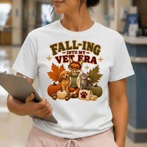 Puede incluir: Una camiseta blanca de manga corta con un estampado gráfico que incluye el texto FALL-ING INTO MY VET ERA. El diseño muestra un zorro con gafas, un cachorro dorado, un gato atigrado, calabazas, hojas de otoño y un pequeño maletín médico.