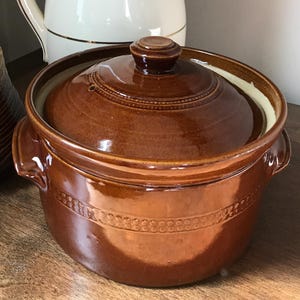 May include: A white ceramic jug with gold trim, a brown glazed ceramic lidded pot with side handles, and a smaller brown textured lidded jar. The jug contains purple flowers. The items are arranged together as a collection of kitchenware.