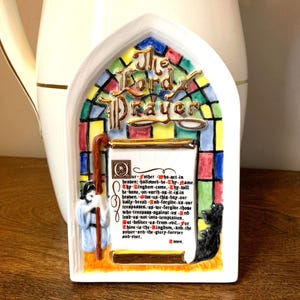 May include: A white ceramic plaque shaped like a gothic arch featuring a stained glass window design in red, blue, green, and yellow. The text The Lord's Prayer is in gold, with the full prayer printed on a central scroll and a figure to the side.