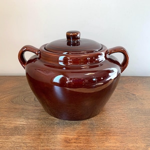 May include: A dark brown glazed ceramic pot with a matching lid and two side handles. The pot features a rounded body and a smooth, reflective surface. The lid has a small, centred knob handle on top, and the entire piece is finished in a deep, uniform brown colour.