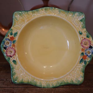 May include: A square ceramic bowl with a pale yellow centre and a wide, textured rim featuring a green lattice pattern. The rim is decorated with hand-painted floral clusters in shades of pink, blue, orange, and green on opposite sides.