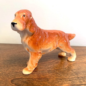 May include: A glossy ceramic figurine of a standing dog with a reddish-brown coat and long ears. The canine figure features a smooth, reflective surface, a black nose, and light-coloured paws, captured in a side profile view with its head turned forward.