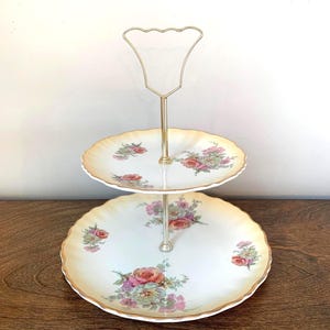May include: A two-tier serving stand featuring white porcelain plates with scalloped edges, a pale yellow rim, and floral patterns in pink, green, and blue. The stand includes a central silver-toned metal rod with a decorative loop handle at the top.