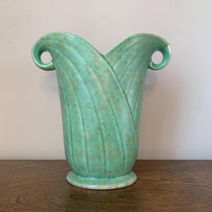 May include: A turquoise ceramic vase with a mottled brown finish, featuring a flared, scalloped rim and two integrated scroll-shaped handles on the sides. The body of the vase has vertical, curved ridges that extend from the base to the top.