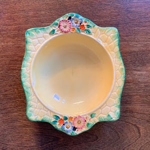 May include: A square ceramic bowl with a pale yellow interior and a textured, cream-coloured rim featuring a green border. The top and bottom edges are decorated with raised, colourful floral motifs in shades of pink, blue, and orange.