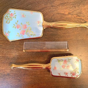 May include: A vanity set consisting of a hand mirror, a hairbrush, and a comb. The mirror and brush feature a light blue backing with a pink floral and gold ribbon pattern, complemented by polished gold-tone handles and trim. The comb is clear.