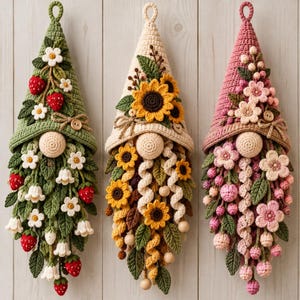 Può includere: Tre ornamenti a forma di gnomo realizzati all'uncinetto con cappelli a punta. Lo gnomo a sinistra ha un cappello verde con fragole e fiori bianchi. Quello centrale ha un cappello color crema con girasoli. Quello a destra ha un cappello rosa.