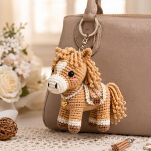 Puede incluir: Un llavero de caballo tejido a ganchillo con cuerpo color canela, hocico blanco y crin rizada. El caballo está adornado con un collar de perlas, un dije de corazón dorado y una pequeña silla de montar con bordado floral y un botón marrón.
