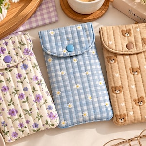 Pode incluir: Três bolsas retangulares acolchoadas com fecho de aba e botão de pressão. A bolsa da esquerda é creme com flores roxas, a central é xadrez azul e branco com margaridas e a da direita é bege com ursos castanhos e corações brancos.