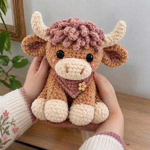 Pode incluir: Um peluche de croché em forma de vaca Highland com corpo castanho-claro, cascos e chifres cor de creme e uma juba encaracolada malva. A vaca usa uma bandana malva com um pequeno botão de flor de madeira e tem olhos de segurança pretos.
