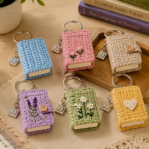 Puede incluir: Seis llaveros rectangulares de ganchillo con forma de libro en azul, rosa, beige, morado, verde y amarillo. Cada uno incluye una anilla metálica, un dije de libro y detalles bordados o botones, como flores, un corazón y un cierre de madera.