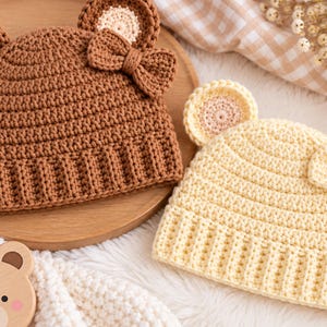 May include: Two crocheted beanies, one brown and one cream, each featuring two rounded bear ears and a small matching bow on the side. The hats have a ribbed brim and a textured stitch pattern, designed with a classic rounded shape for a snug fit.