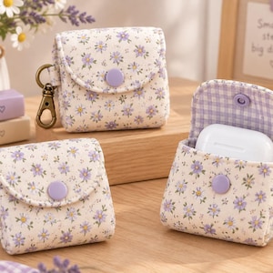 May include: Three fabric earbud cases featuring a white background with a purple floral pattern. Each case has a purple snap button closure and a bronze-toned metal lobster clasp. One case is open, revealing a white earbud charging case inside.