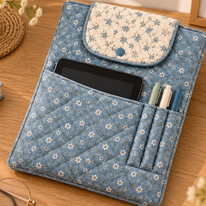 May include: A quilted blue fabric tablet sleeve featuring a white floral pattern, a flap closure with a blue button, an external pocket containing a black tablet, and three vertical slots holding a light green, white, and blue pen.
