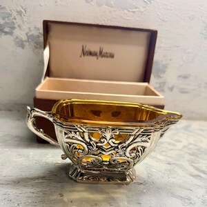 Può includere: Una lattiera con un inserto in vetro ambrato traslucido racchiuso in una cornice decorativa in metallo argentato con intricati motivi a volute e un manico. La lattiera è presentata in una scatola marrone aperta con il logo Neiman Marcus.