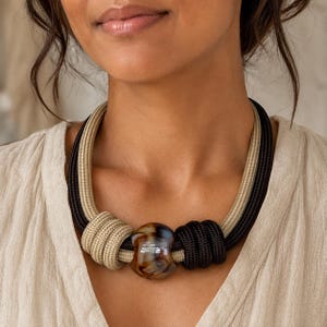 May include: A statement necklace featuring thick, braided rope strands in black and beige. The design includes a central, polished, multi-colored brown and cream marbled bead, with the rope coiled around it on both sides to create a textured look.