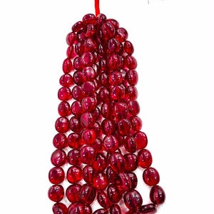 May include: A strand of translucent, deep red, oval-shaped beads arranged in multiple vertical columns. The beads have a smooth, polished finish and are strung together, culminating in a bright red thread tassel at the top of the strand.