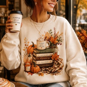 Pode incluir: Uma camisola bege de gola redonda com uma impressão gráfica de uma pilha de livros, abóboras, uma vela e uma caneca com o rótulo Pumpkin Spice Everything. O design inclui folhas de outono, um cachecol aos quadrados e mangas compridas.