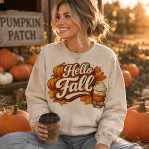 Puede incluir: Una sudadera beige de cuello redondo con un estampado gráfico que incluye el texto Hello Fall, calabazas naranjas, hojas de otoño y una taza de café con nata montada. El diseño está centrado en el pecho de la prenda de manga larga.