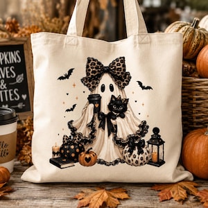 May include: A cream-colored canvas tote bag featuring a graphic of a ghost wearing a leopard print bow, holding a coffee cup and a black cat. The design includes small bats, pumpkins, a lantern, a candle, and a book, all in a fall-themed aesthetic.