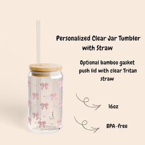 Könnte beinhalten: Ein transparenter Glasbecher mit Bambusdeckel und durchsichtigem Strohhalm. Der Becher ist mit einem Muster aus kleinen rosa Schleifen und dem Namen Emma bedruckt. Der Text lautet: Personalized Clear Jar Tumbler with Straw, Optional bamboo gasket push lid with clear Tritan straw, 16oz, und BPA-free.