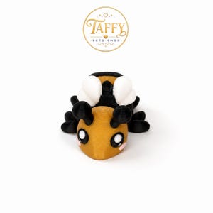 May include: A small bee figurine featuring a golden-yellow head with black antennae, large black and white eyes, and pink cheek accents. The body is black with white wings and multiple black legs, all rendered in a textured, layered 3D printed finish.