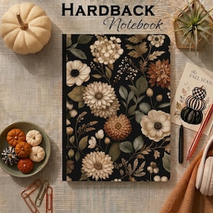 May include: A hardback notebook with a black cover featuring a floral pattern of beige, cream, and brown dahlias and leaves. The text "HARDBACK Notebook" is printed at the top. The notebook is surrounded by small pumpkins, a plant, and stationery.