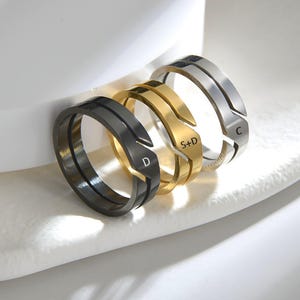 Könnte beinhalten: Drei Metallringe in Schwarz, Gold und Silber, jeweils mit einem geteilten Banddesign. Der schwarze Ring ist mit dem Buchstaben D, der goldene Ring mit S+D und der silberne Ring mit dem Buchstaben C auf ihren jeweiligen flachen Oberflächen graviert.