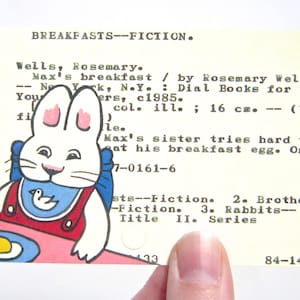 Puede incluir: Una tarjeta de biblioteca vintage con una ilustración de un conejo en una mesa con un huevo frito. La tarjeta incluye el título "Max's breakfast" y el nombre de la autora, Rosemary Wells. La tarjeta mide 16 cm y está sujeta por una persona.