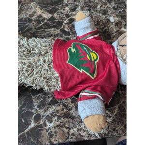 Puede incluir: Un peluche de pelaje marrón claro desgreñado que lleva una camiseta roja. La camiseta presenta un logotipo gráfico verde y amarillo de una cabeza de animal estilizada. El juguete tiene mangas gris claro y patas de tela color canela.
