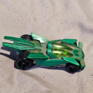 May include: A green toy race car with a futuristic design, featuring sharp angular body panels, two translucent lime green cockpit windows, and four black plastic wheels. The vehicle has an elongated rear section with dual fins.