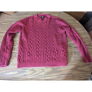 May include: A long-sleeved crew neck sweater in a deep red color. The garment features a vertical cable knit pattern throughout the front and sleeves, with a ribbed knit texture at the collar, cuffs, and hemline. A small dark brand tag is visible.
