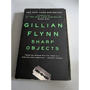 May include: A paperback book titled Sharp Objects by Gillian Flynn with a black cover and bright green text. The cover features the text New York Times Bestseller, the author name, and a graphic of a silver razor blade at the bottom centre.