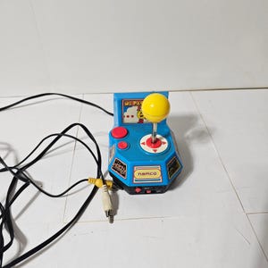 Könnte beinhalten: Ein blauer Plug-and-Play Ms. Pac-Man Videospiel-Controller mit einem gelben Joystick, zwei roten Tasten und einem Namco-Logo. Das Gerät verfügt über ein langes schwarzes Kabel mit gelben und weißen Cinch-Anschlüssen für den Fernseher.