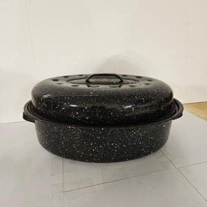 May include: An oval-shaped black roasting pan with a speckled white enamel finish. The roaster features a matching lid with a central handle and integrated side handles on the base, providing a classic look for cooking large meals in the oven.
