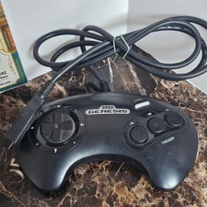 Puede incluir: Un mando de videojuegos Sega Genesis negro con una cruceta circular a la izquierda, tres botones de acción redondos etiquetados como A, B y C a la derecha, un botón Start blanco y un cable negro largo con un conector de enchufe.