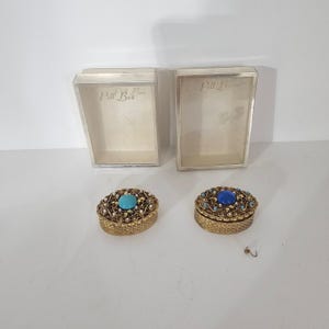 May include: Two oval gold-tone metal pill boxes with intricate filigree patterns, each featuring a central cabochon stone—one turquoise and one dark blue—surrounded by small white accents. Two rectangular clear plastic cases labeled Pill Box.
