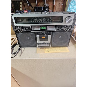 May include: A vintage JVC Biphonic portable stereo boombox featuring a silver and black chassis, dual speakers, a central cassette deck, analogue radio tuning dials, and illuminated VU meters with green accents, including a power cord and manual.